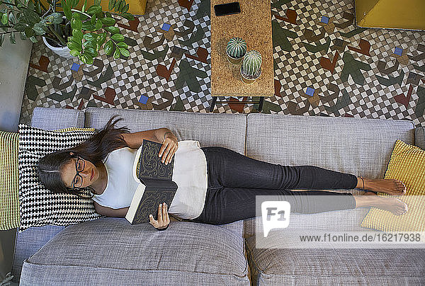 Frau liest ein Buch  während sie zu Hause auf dem Sofa liegt