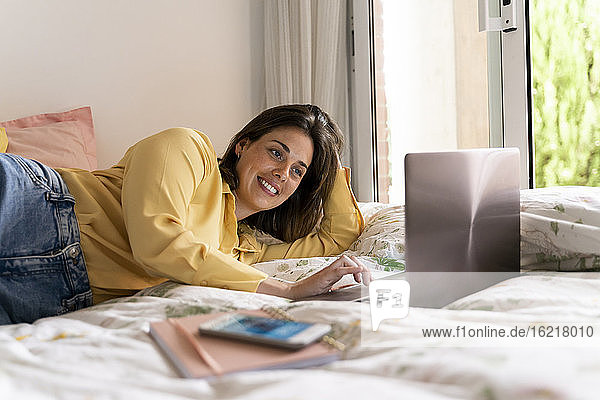 Smiling woman using laptop while lying in bedroom