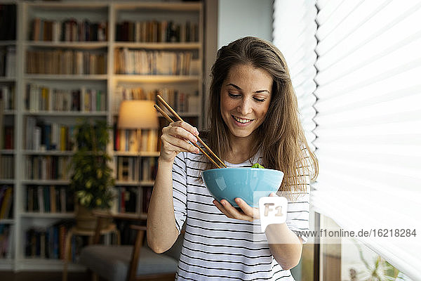 Lächelnde junge Frau  die zu Hause am Fenster sitzt und isst