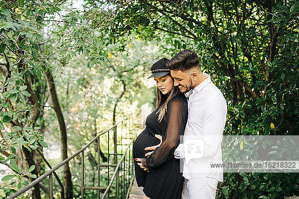 Smiling man embracing and touching belly of woman while standing in park
