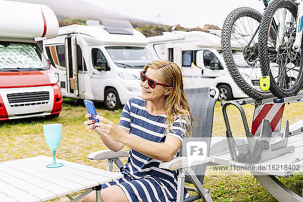 Entspannte Frau  die neben einem Wohnmobil sitzt  ihren Urlaub genießt und ihr Smartphone benutzt