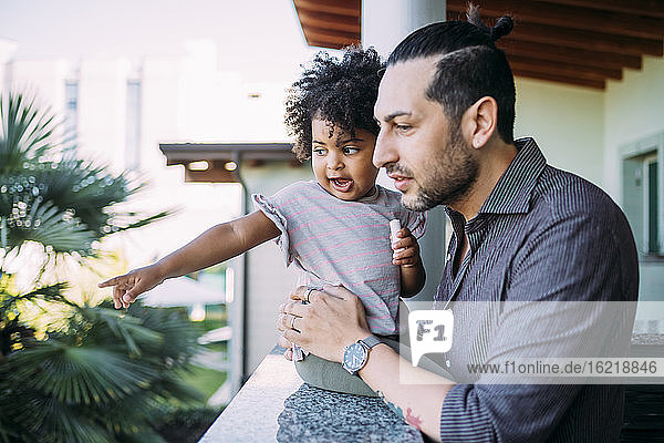 Vater hält kleines Mädchen  das auf eine Stützmauer zeigt  während er auf einem Balkon steht