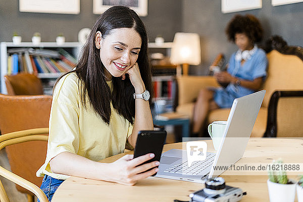 Happy woman using smart phone at cafe table with friend in background