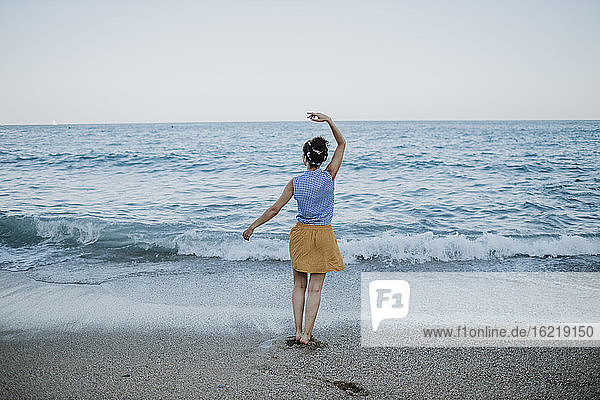Mid adult woman with arm raised standing at shore against clear sky during sunset