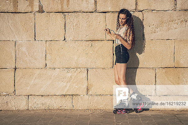 Junge Frau fährt Inline-Skates und lehnt sich mit ihrem Smartphone an eine Wand