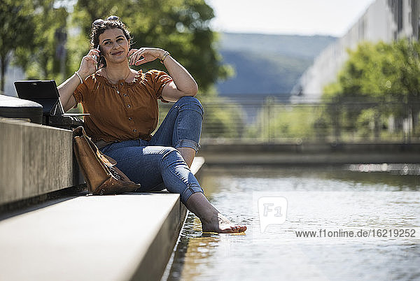 Lächelnde Frau  die über ihr Handy spricht  während sie an einem sonnigen Tag am Teich im Park sitzt