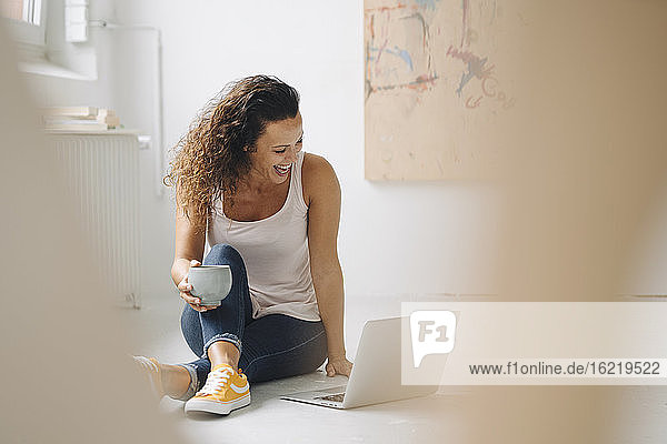 Cheerful woman holding coffee mug while using laptop on floor at home