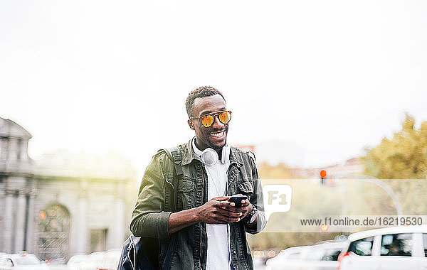 Lächelnder Mann mit Sonnenbrille  der ein Smartphone benutzt  während er vor einem klaren Himmel in der Stadt steht