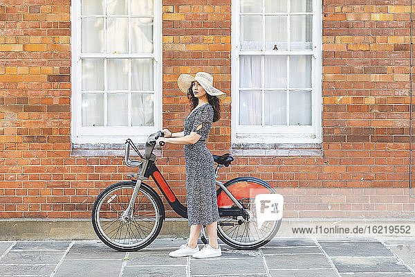 Junge Frau mit Hut und Fahrrad steht auf der Straße neben einem Gebäude in der Stadt