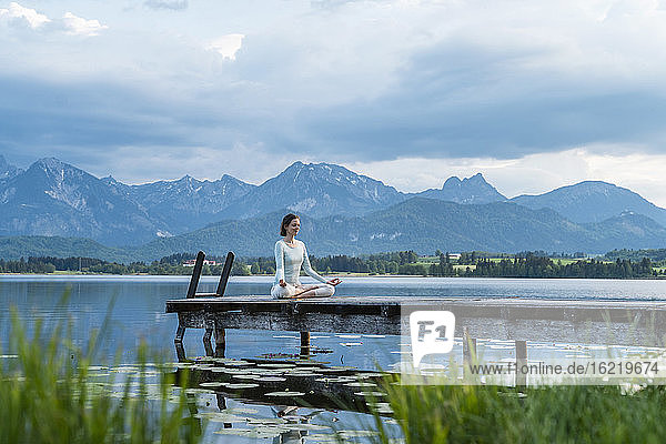 Mittlere erwachsene Frau meditiert auf einem Steg über einem See vor bewölktem Himmel