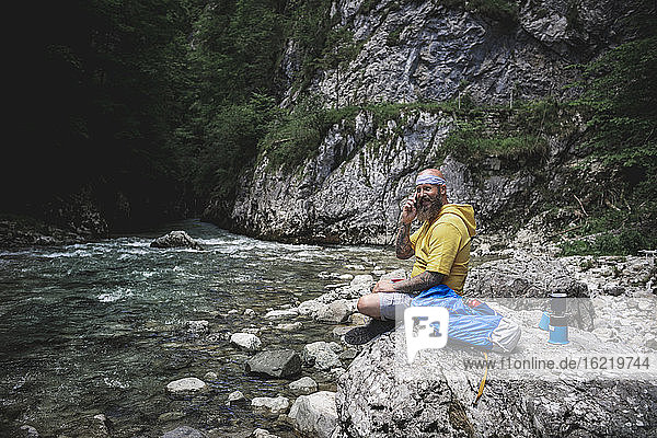 Hiker with full beard and yellow hoodie sitting on stone next to river and using smartphone