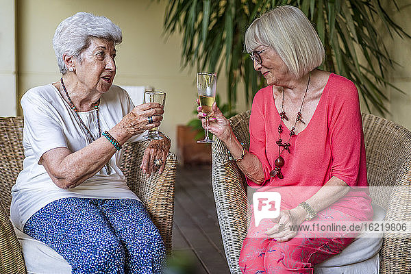 Ältere Freundinnen stoßen mit Sektflöten an  während sie auf Stühlen im Innenhof sitzen