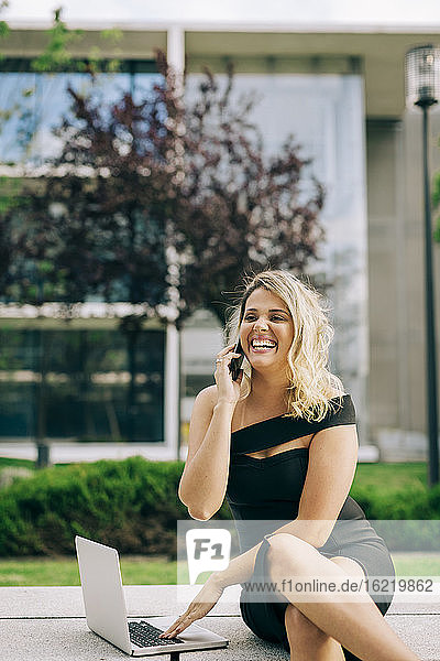 Fröhliche Geschäftsfrau  die mit dem Handy telefoniert und einen Laptop im Freien benutzt