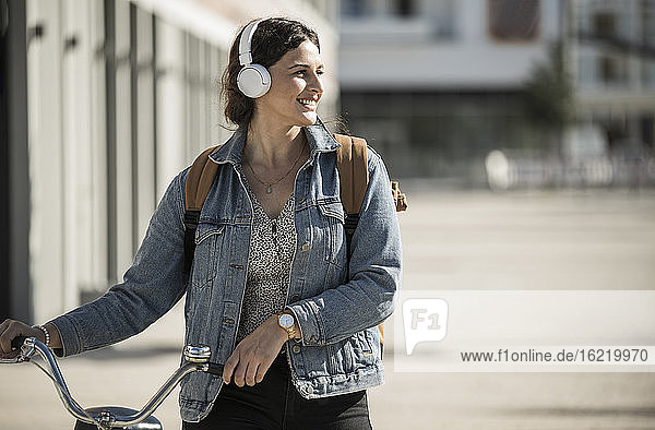 Smiling woman listening music through headphones while walking with bicycle in city