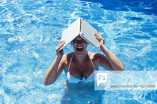 Fröhliche Frau mit Laptop auf dem Kopf beim Schwimmen im Pool