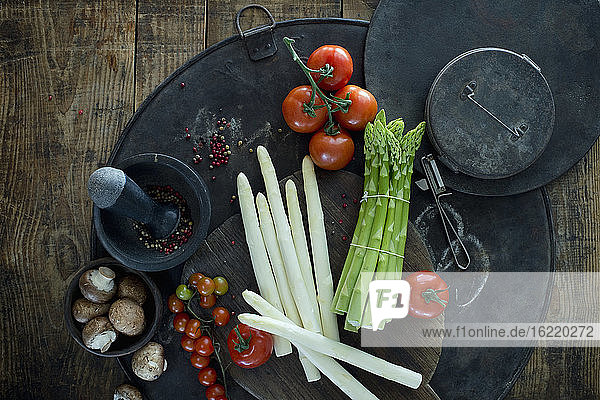 Mörser und Stößel  Schäler  Spargelstangen  Pfefferkörner  Tomaten und Schüssel mit Pilzen auf rustikalem Backblech