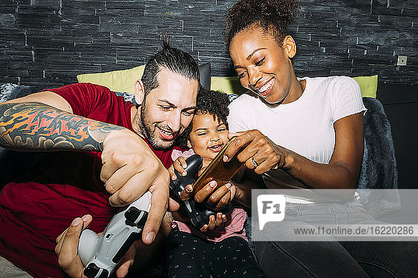 Fröhliche Mutter  die ein Smartphone benutzt  während Vater und Tochter auf dem Sofa zu Hause ein Videospiel spielen