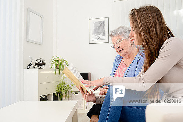 Cheerful young woman reading book for old senior woman at home