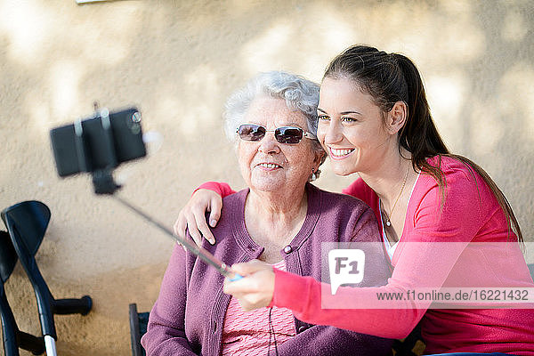 Fröhliche junge Frau macht ein Selfie mit einer älteren Frau in einem Garten eines Seniorenheims.