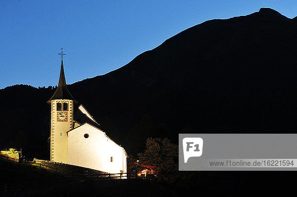 Schweiz  Kanton Wallis  Binntal  die Kirche auf der Höhe des Dorfes  die bei Einbruch der Dunkelheit beleuchtet ist und sich von den Bergen abhebt.