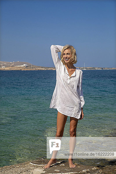 Eine schöne junge Frau im weißen Hemd steht vor dem Meer in der Sonne.