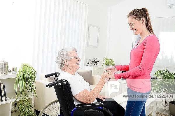 Fröhliche junge Frau  die sich zu Hause um eine ältere Frau im Rollstuhl kümmert