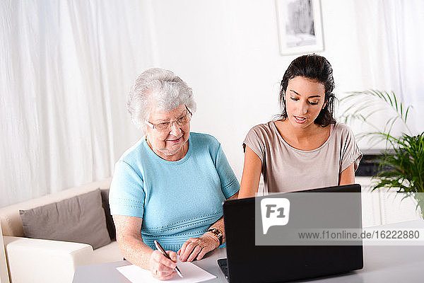Fröhliche junge Frau  die einer älteren Person bei der Nutzung eines Laptops für die Internetsuche und E-Mail hilft