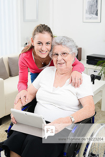 Fröhliches junges Mädchen  das mit einem Tablet-Computer im Internet spielt und Zeit mit einer alten Seniorin im Rollstuhl verbringt