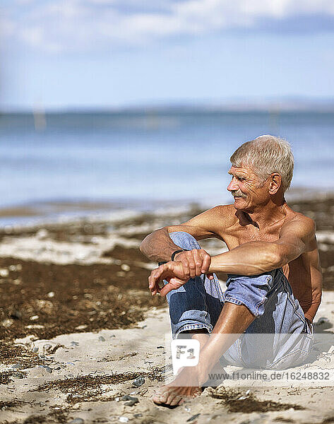 Älterer Mann in Jeans sitzt am Sandstrand