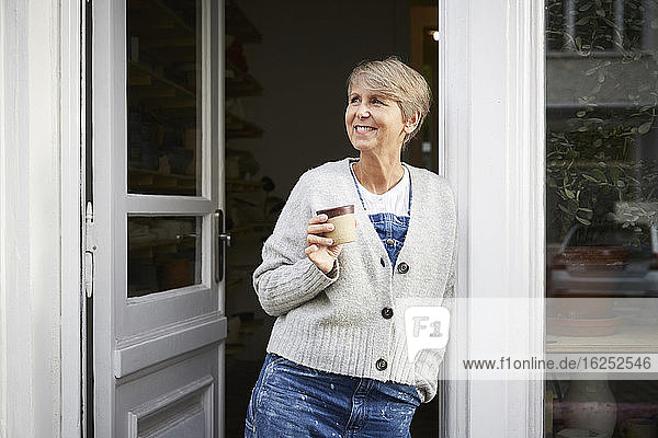 Lächelnde reife Frau trinkt Kaffee am Eingang des Kunstateliers
