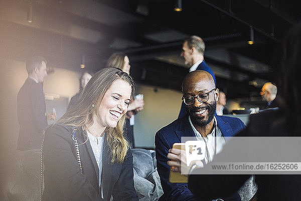 Smiling male and female coworkers looking at mobile phone while sitting with entrepreneur in office