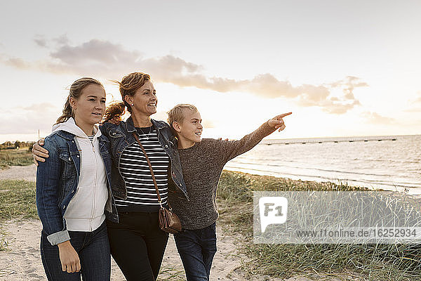 Lächelnder Sohn zeigt  während Mutter und Tochter am Strand vor bewölktem Himmel stehen