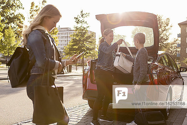 Seitenansicht eines Teenagers beim Telefonieren  während Mutter und Sohn die Tasche im Kofferraum des Autos aufbewahren