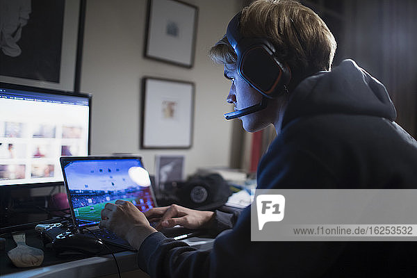 Teenage boy with headphones playing video game at laptop in dark room