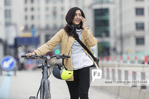 Smiling woman with bicycle talking on smart phone in city