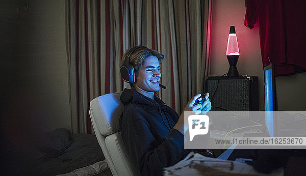 Smiling teenage boy playing video game at computer in dark bedroom