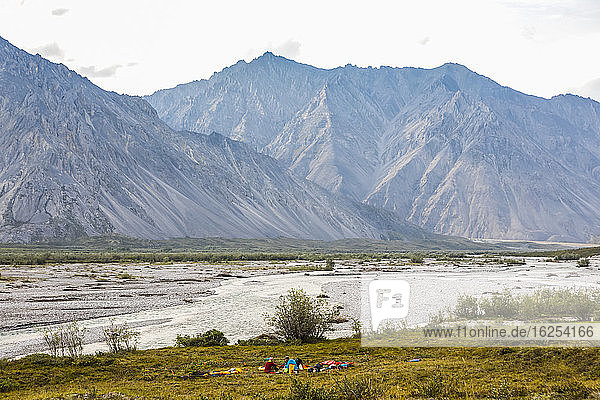 Caucasian man and woman in the cooking area of camp  surrounded by their bear fence  are dwarfed by the landscape  with the Marsh Fork river flowing by and gigantic Brooks Range mountains rising up in the background in summer; Alaska  United States of America