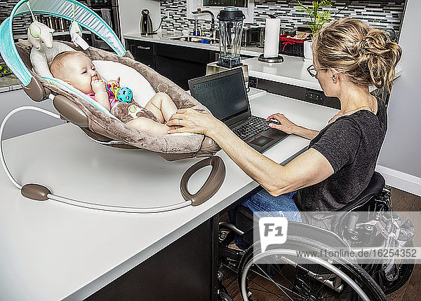 A young mom with a spinal cord injury looks after her newborn baby while working on her computer in the kitchen; Edmonton  Alberta  Canada