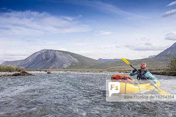 Caucasian woman in her 40's smiles as she digs deep with her paddle into the river  guiding her yellow packraft down the crystal clear Marsh Fork River  in the Brooks Range  on a sunny summer day  Arctic National Wildlife Refuge  Alaska  while another boat is in the background way behind her; Alaska  United States of America