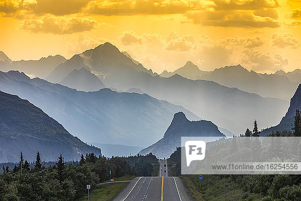 Glenn Highway teilt das Bild  umgeben von Fichtenwald und Gestrüpp im südlichen Zentralalaska  bei Sonnenuntergang  mit Autos in der Ferne  die in einer verrauchten  dunstigen Sommernacht auf Lion's Head und die Chugach-Berge blicken; Chickaloon  Alaska  Vereinigte Staaten von Amerika