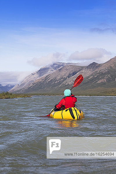Woman wearing red drysuit and pfd  paddling yellow packraft boat down the Canning river through the Brooks Range  Arctic National Wildlife Refuge; Alaska  United States of America