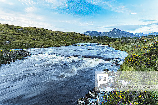 Small rapids on a river flowing through Connemara with a mountain in the background on a blue sky day; Connemara  County Galway  Ireland