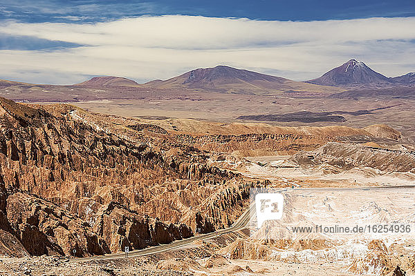 Die Straße führt hinunter in ein hochgelegenes Wüstental mit einzigartigen Felsformationen auf der linken Seite und einem Vulkangipfel in der Ferne; San Pedro de Atacama  Antofagasta  Chile