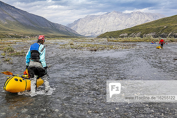 Caucasian woman wearing blue drysuit wades downriver guiding her yellow packraft through a shallow braided section of the Marsh Fork river  Brooks Range  Alaska  on a sunny  summer day. A woman in red drysuit with yellow boat drags hers downstream in the background and man wearing orange  paddles his blue boat in very background  Arctic National Wildlife Refuge  Alaska; Alaska  United States of America