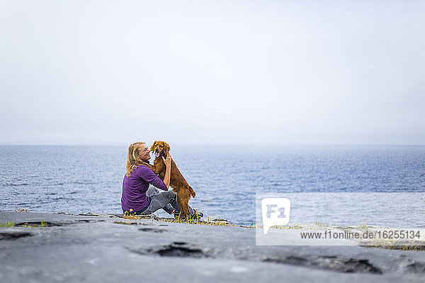 Woman embracing dog on a rocky ground overlooking the sea on a cloudy day; Fanore  County Clare  Ireland