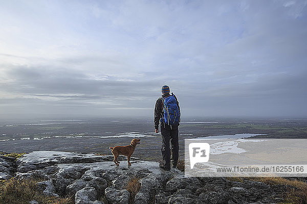 Man with backpack and dog standing on a cliff looking out at the lakes in the Burren National Park; County Clare  Ireland