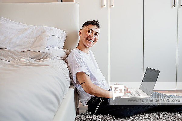 Junge Frau mit rasiertem Kopf sitzt im Schlafzimmer  benutzt einen Laptop und lächelt in die Kamera.