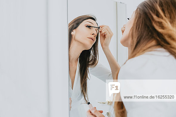 Frau mit braunem Haar steht vor einem Spiegel und trägt Wimperntusche auf.
