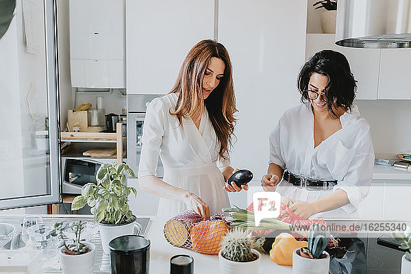 Zwei lächelnde Frauen mit braunem Haar stehen in einer Küche und nehmen Gemüse aus dem Einkaufsnetz.