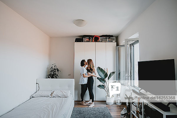 Zwei Frauen mit braunem Haar stehen in einer Wohnung  umarmen und küssen sich.